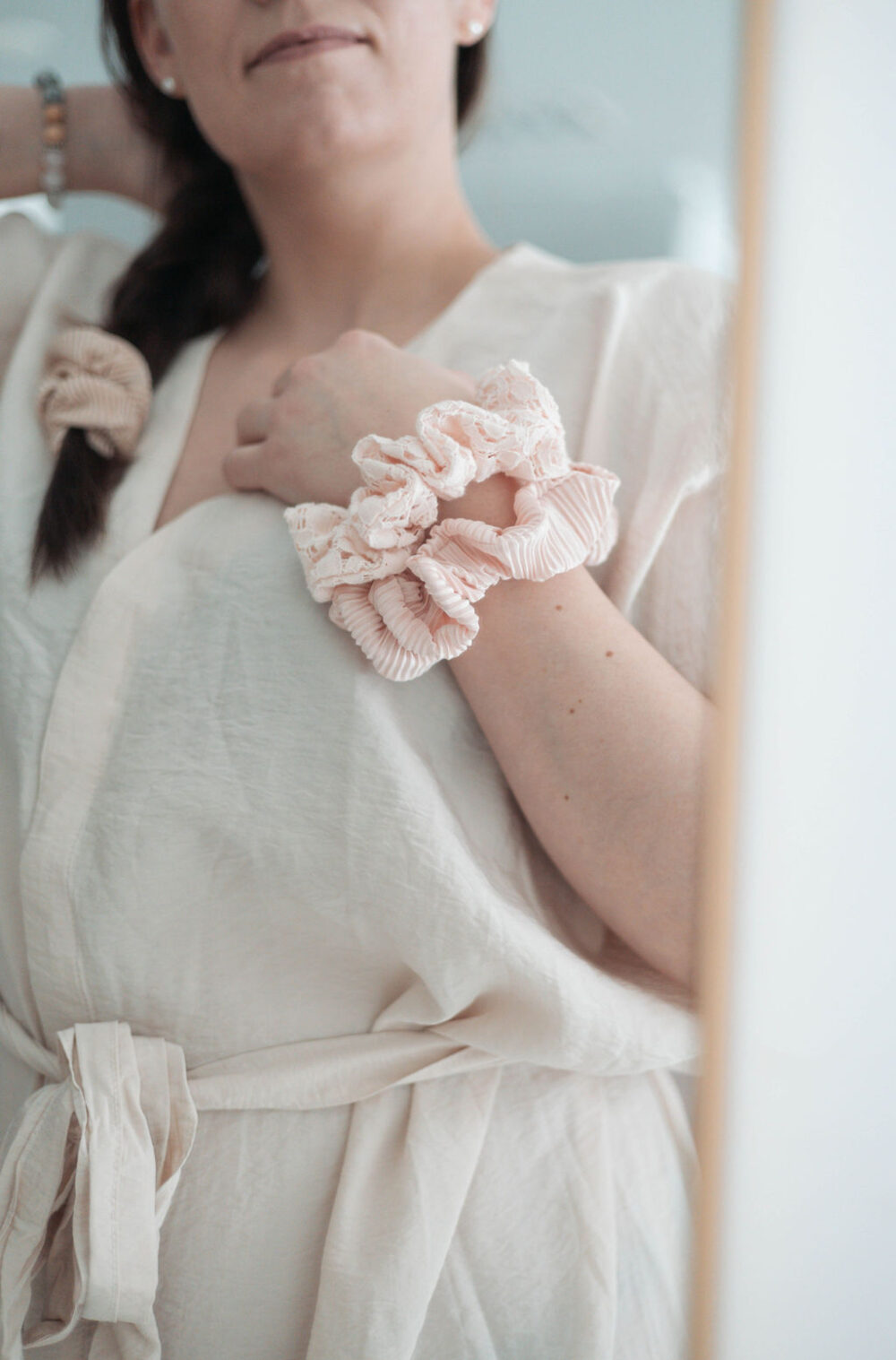 Nainen valkoinen mekko päällään katsoo itseään peilistä. Naisen käsi on hänen rinnuksellaan, ranteessa on kaksi vaalean roosan väristä hiusdonitsia.